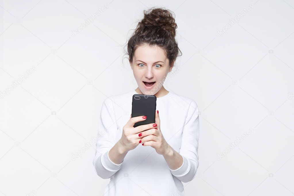 Excited happy stylish hipster lady reads news online, uses free internet connection on modern cell phone, finds out big sales. Surprised female model receives pleasant message about winning a prize.