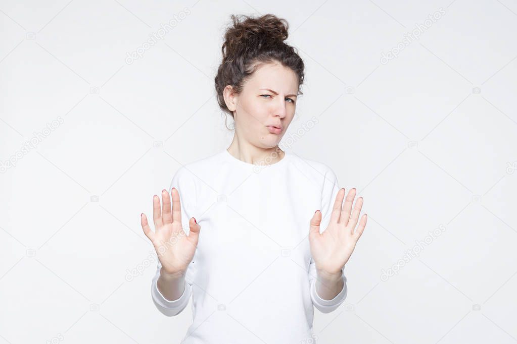 Body language. Disgusted stressed out beautiful young woman with hair knot posing at studio wall, keeping hands in stop gesture, grimacing, trying to defend herself as if saying: Stay away from me.