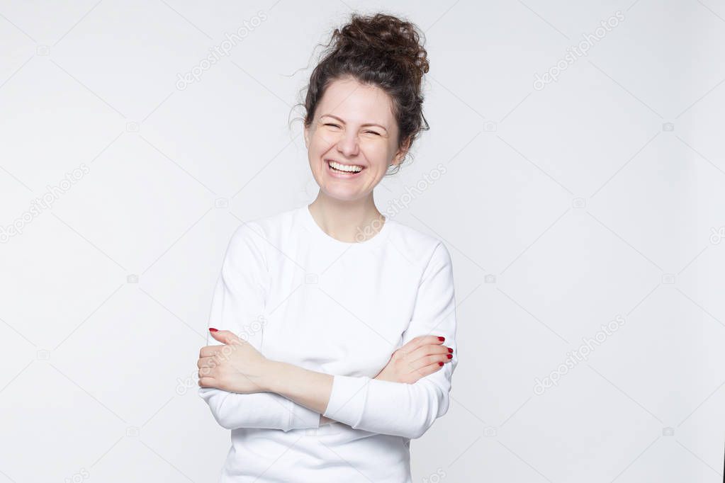 Smiling positive female with attractive look, wearing long sleeved white T-shirt, posing against blank wall. Happy woman with hair bun showing positive emotions after receiving pleasant compliment.