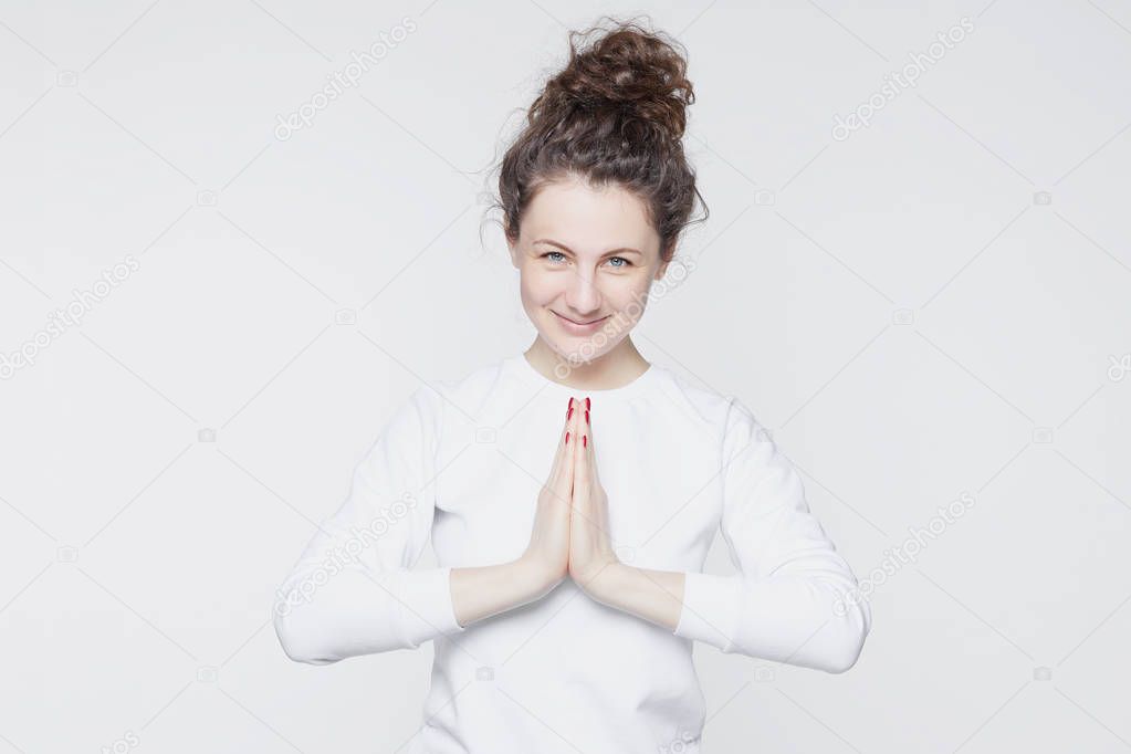 People and lifestyle concept. Beautiful young Caucasian woman with curly bun hairstyles, dressed in casual white t-shirt, smiling happily, looking at camera, holding hands in prayer or namaste mudra.