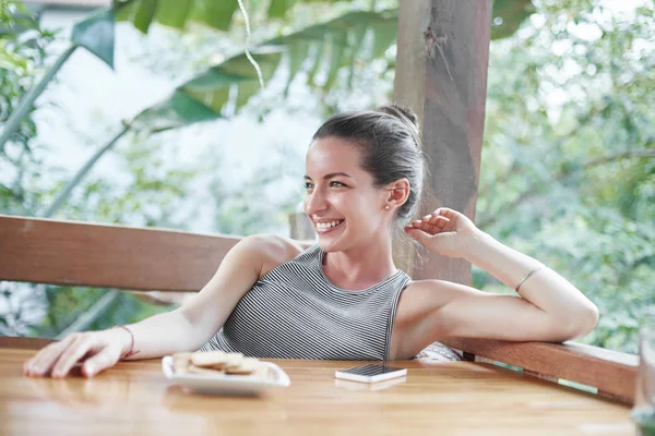 Adorable happy smiling Caucasian female has excited expression, poses at cafe alone, being lucky to travel, dressed casually, has good recreation. Relaxed mixed race woman enjoys leisure time outdoor.