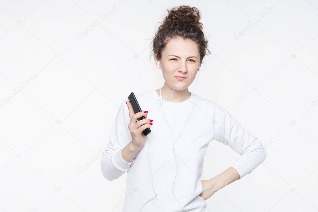 Playful attractive coquette woman raises eyebrows and pouts lips, listens to new album of famous band, using earphones and mobile phone, stands over white background with copy space for your text.