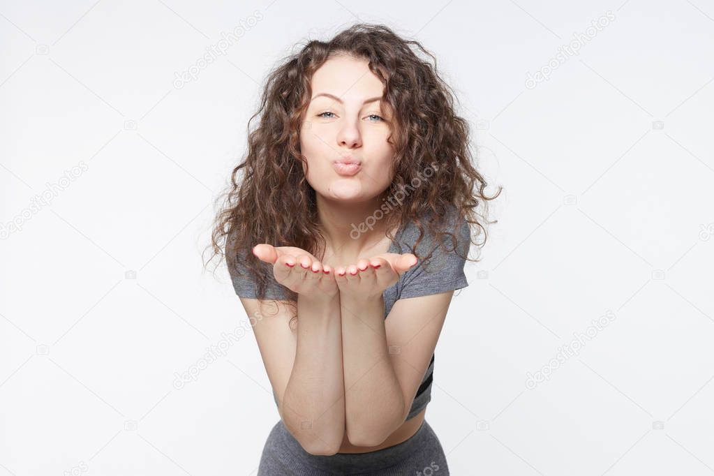 Young attractive European woman posing with kiss on her lips, wearing trendy dress, having flirty look, feeling confident and beautiful. Charming grey eyed lady with Afro hair having fun indoors.