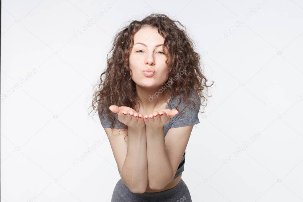Young attractive European woman posing with kiss on her lips, wearing trendy dress, having flirty look, feeling confident and beautiful. Charming grey eyed lady with Afro hair having fun indoors.