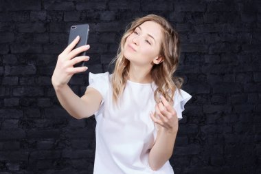 Cep telefonu kullanarak, bir öpücük gönderme otoportre yapma beyaz elbise güzel sarışın Bayan portresi. Genç kadın sevimli yüzü siyah tuğla karşı fotoğraf çekme ile stüdyo arka plan duvar. 