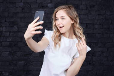Kullanımının güzel dalgalı saçlı Bayan giyen beyaz kadınsı elbise yapma kendi kendine portre portre cep telefonu, açılır ağız. Genç sarışın kadın mavi gözlü siyah tuğla duvara fotoğraf çekmek. 