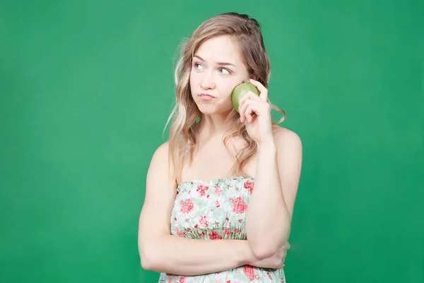 Healthy food and lifestyle concept. Pretty female eats apple, looks for new diet ,being glad to loose weight. Blonde woman posing on green wall background making new video blog for social networks .