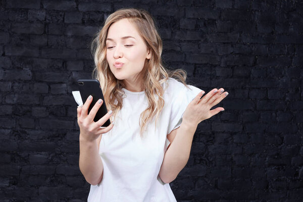 Close up shot of young pretty  kissing blonde lady holding smartphone digital camera with  hand and taking selfie portrait, enjoying youth and beauty, standing indoors against black loft background.