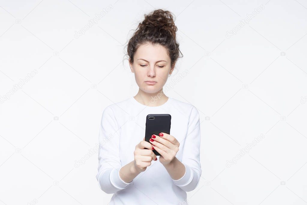 Displeased upset female curves lip, being unhappy to receive bill from banking service on mobile phone, has no money to pay, feels frustrated and disappointed, isolated over white background.