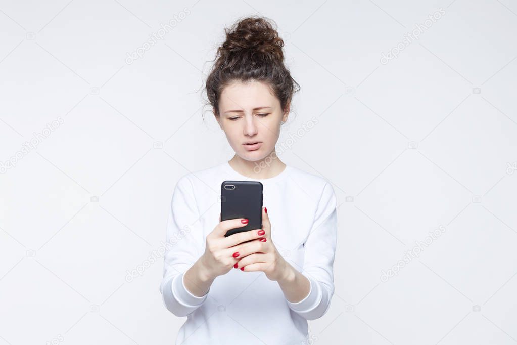 Close-up portrait of young American woman with curly hair in bun wearing casual T-shirt communicating over smartphone video, having sorrowful,sad expression. Human emotions and feelings concept.