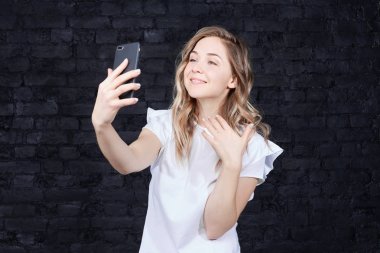 İnsan yüzü ifade ve duygular. Ayna kullanarak cep telefonu tutarak ve kameraya, arıyorum memnun sarışın genç kadın görüntüsünü kırpılmış. Oldukça selfie tuğla duvara alarak kendini hayran.