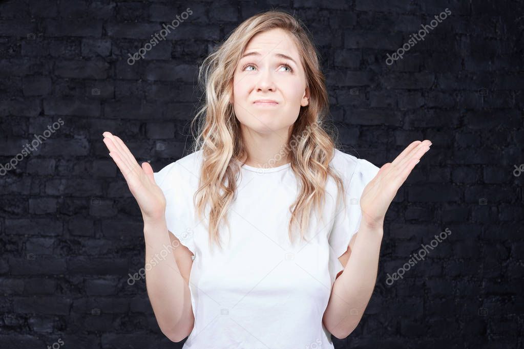 Are you kidding me? Wtf! Close up portrait of cute disappointed woman with big blue eyes, long wavy hairstyle looking up, talking to God, expressing negative, isolated on black brick wall background
