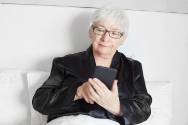 Yaşlı Olgun kadın siyah ipek pijama resimleri online Elektronik Gadget'ı kullanarak Internet üzerinde görüntüleme sabah yatakta rahatlatıcı. Kırpılan üst düzey beyaz kadın akıllı telefon e-kitap okuma görünümünü.