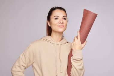 Güçlü, kendine güvenen genç bir Amerikalı kadın kameraya saygınlık ve güçle bakıyor, yoga minderini elinde tutuyor, üstünkörü bir kapüşonlu giyiyor, gri duvarda reklam için fotokopi alanı ile poz veriyor. Sağlık ve güzellik.