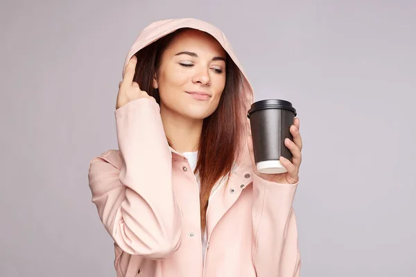 Close up shot of pleased lovely dark skinned young woman wears raincoat with hood, drinks hot coffee during stormy weather, closes eyes, enjoys good moment, has healthy look, isolated over grey wall.