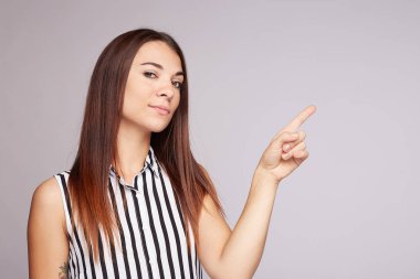 Horizontal shot of attractive tan skinned woman with long straight shiny hair, points index finger on copy space, advertises object on blank wall, looking at camera, dressed in striped blouse,isolated 