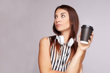 Studio shot of thoughtful brunette woman with dark long straight hair, enjoys drinking takeaway coffee, wears black white blouse, headphones on neck,has natural beauty, isolated over grey background. 
