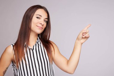 Horizontal shot of joyful pleased good looking youngster points at upper right corner with index finger, invites you go there, dressed in casual blouse, poses against grey background, blank copy space
