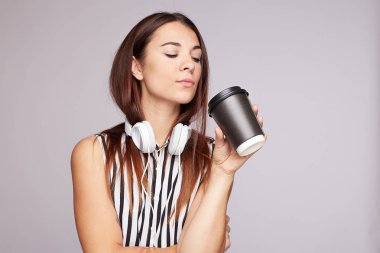 Pretty tan skinned lady enjoys deep music in stereo headphones, holds disposable cup of drink, wears silk blouse, spends free time listening favorite songs, models over grey studio wall, blank space.