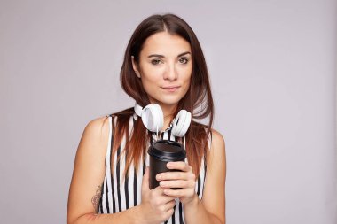 Studio portrait of glad female student has coffee break after lectures, listens audio book in headphones, enjoys record from website, holds disposable cup with hot beverage, isolated indoors on grey.
