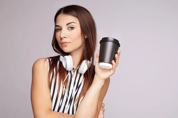 音楽の時間だ自信に満ちた日焼け肌の女性は使い捨てカップに行くためにコーヒーを保持しています ヘッドフォンで快適なメロディーを聞きに行きます カメラで浮気見えます カジュアルストライプブラウスを身に着けています スタジオでポーズ — ストック写真