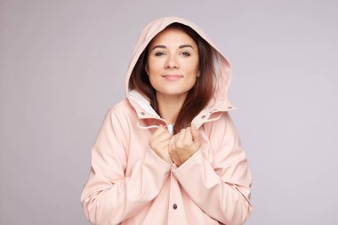 Hoş, şirin, melez bir kadının portresi soluk pembe bir başlık giyiyor, su geçirmez bir yağmurluk giyiyor, ellerini yakasında tutuyor, olumlu duygular ifade ediyor, doğal güzelliği var, kapalı alanda gri stüdyo arka planında poz veriyor..