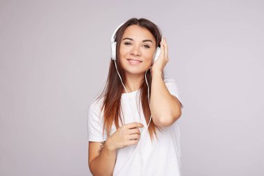 Çekici kızıl saçlı kadın, mutlu görünüş, büyüleyici gülümseme, kulaklıkla el ele tutuşma, modern cihazla en sevdiği müziği dinleme, beyaz tişört giyme, kapalı alanda gri pozlar verme. İnsanlar, teknoloji, boş zaman.