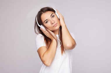 Gündelik giysiler içinde çekici bir kadın, nazik ve kurnaz bir şekilde gülümsüyor, modern cep telefonu kullanıyor, kulaklıkla müzik dinliyor, en sevdiği parçayı seçiyor, gri arka planda poz veriyor. Millet, aktif yaşam biçimi içeriği.