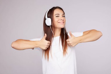 Mutlu, bronz tenli, güzel bir kadın iki başparmağıyla da güzel bir jest yapar, geniş bir şekilde gülümser, kameraya poz verir, favori müzikten hoşlanır, modern kulaklıklar kullanır, gri duvarda fotokopi alanı olan kapalı bir yerde poz verir..