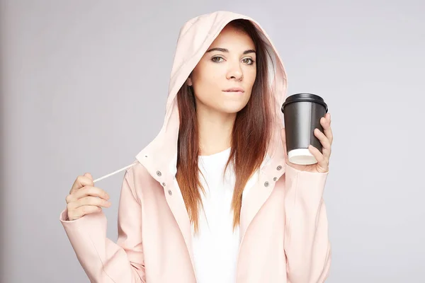 Beautiful young Caucasian female student has natural beauty with no make up, bites lip in hood of raincoat, drinks takeaway coffee, ready for studying, looks confidently, gets knowledge at university.