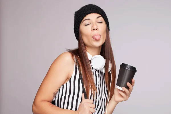 Horizontal shot of glad teenage female licks lips with tongue, holds disposable cup of aromatic coffee, takes break after analyzing piechart, wears striped blouse, listens audio book, isolated on grey