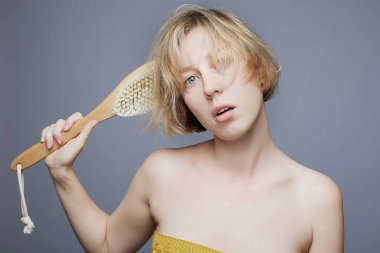 Kameraya bakan tuhaf, suratını buruşturan, kocaman mavi gözleri olan, çıplak omuzları olan, kısa sarı saçlı, kocaman tahta masaj fırçalı, güzellik tedavi konsepti.