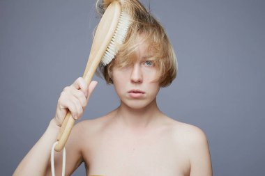 Şehvetli bir kadının kameraya masum, derin mavi gözleriyle baktığı, yumuşak dudakları açık, çıplak vücudu olan, kısa sarı saçlara kuru masaj için ahşap fırça tutan, güzellik terapisi konsepti