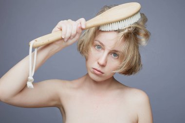 Şehvetli masum bir kadının, büyük mavi gözleriyle, çıplak bedeniyle kameraya bakarken, kısa sarı saçlara kuru masaj için ahşap fırça tutarken, güzellik muayenesi yaparken, gri stüdyo duvarında poz verirken..