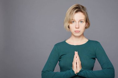 Koyu mavi gözlü, umut verici bir ifade takınan, yalvaran ve dua eden, avuçlarını birbirine bastıran, bir şey için endişelenen, ciddi bakışları olan, yeşil süveter giyen, gri stüdyo duvarına izole edilmiş genç bir kadın.. 