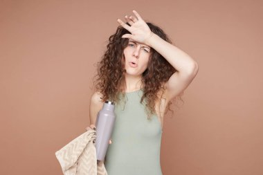 Spor ve aktif yaşam tarzı konsepti. Spor kıyafetleri içinde sağlıklı, zayıf bir kadın, antrenmandan yorgun düşmüş, çelik şişeden su içen, bej renkli stüdyo duvarına karşı duran, boş bir alan..