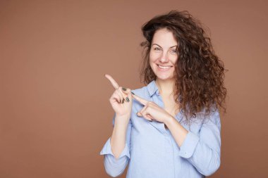 Çekici gülümseyen genç bir kadın sizi oraya davet ediyor, güzel bir kafe tavsiye ediyor, işaret parmağı sol tarafta, mavi pamuklu gömlek giyiyor, bej duvar arkasında izole edilmiş, yönünü gösteriyor..