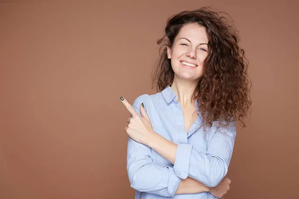 Joyful attractive Caucasian curly haired female with face happy expression, looks at camera positively aside, points aside at beige wall with blank copy space for advertisement or promotional content.