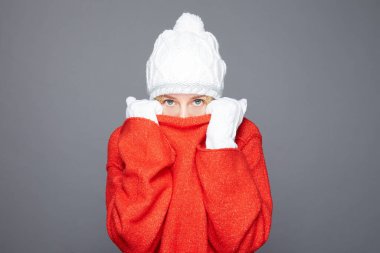 Ne kadar serin. Şaşkın mavi gözlü kadın kendini kucaklıyor, soğuktan titriyor, rüzgarlı havada yürüyor, dişlerini sallıyor ve sıkıyor, kırmızı kazak giymiş, montsuz duruyor, gri stüdyo duvarında izole edilmiş..