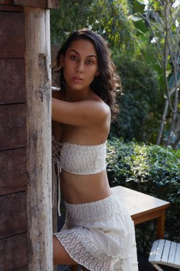 Esmer tenli, çekici, melez bir kadın modelin dikey portresi keten bir bluz giyiyor, kameraya bakıyor, doğal bahçe arka planında poz veriyor. Moda, güzellik, tarz kavramı.