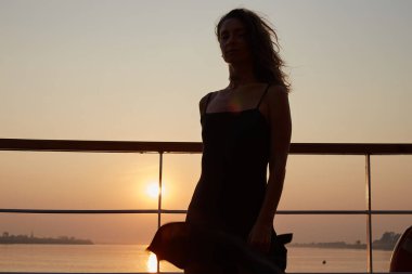 Uzun siyah elbiseli, çekici, otantik beyaz kadın, yolcu gemisinde poz veriyor, mutlu ve kendinden emin hissediyor, muhteşem nehir manzaralı resimli günbatımının keyfini çıkarıyor..