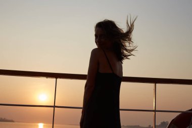 Uzun siyah elbiseli, çekici, otantik beyaz kadın, yolcu gemisinde poz veriyor, mutlu ve kendinden emin hissediyor, muhteşem nehir manzaralı resimli günbatımının keyfini çıkarıyor..