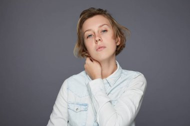 Üzgün, mutsuz, genç bir kadın kendini yalnız hissediyor, boynuna dokunuyor ve düz bakıyor, kameraya bakıyor, çok yorgun, mavi kot gömlek giyiyor, gri stüdyo duvarına karşı poz veriyor. Kötü duygular kavramı.
