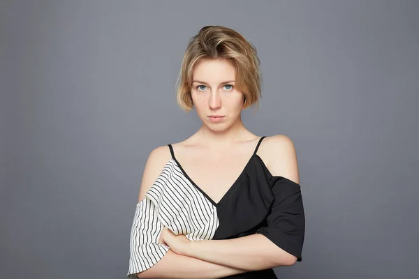 Annoyed pissed female crosses hands over chest,feels insulted, frowns and sulks, waits for explaining situation, looks from eyebrows, wears festive blouse, stands over grey studio wall background.