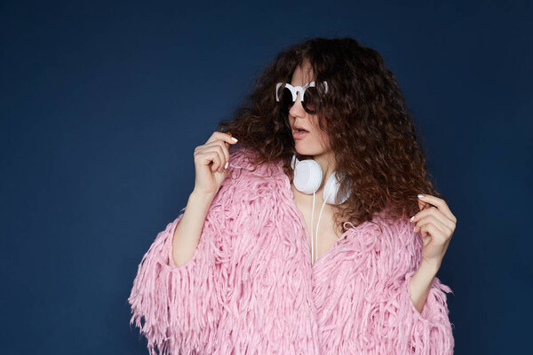 Close up shot of confident posh American female model wears trendy sunglasses, dressed in pink shaggy jacket, prepares for date, isolated on ultramarine wall, has fashionable look, listening to audio.