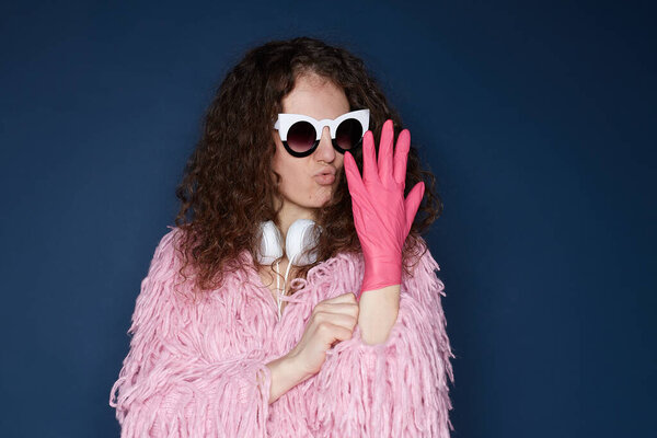 Horizontal portrait of bushy haired Caucasian lady wearing funky pink jacket, fashionable spectacles, headphones on neck, dressing up protective gloves, taking care of herself even prepare home party.