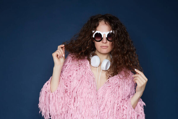 Studio shot of stylish curly headed woman in fashionable pink shaggy jacket, wearing headphones, sunglasses, touching gorgeous hair, isolated on ultramarine wall. People, fashion, luxury concept.