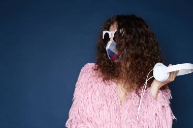 Ev partisi konsepti. Çekici kıvırcık saçlı, koruyucu renkli bir maske takan, tüylü ceket takan, güneş gözlüğü takan, kulaklık takan, ultramariyede izole edilmiş bir kadın..