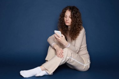 Bin yıllık düşünceli kadın kıvırcık saçlı, yerde oturuyor, telefonunu tutuyor, takipçilerini kontrol ediyor, mesajlar yolluyor, ciddi yüz ifadesi var, mavi arka planda izole edilmiş, boş bir alan..