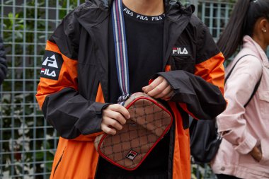 Fila moda defilesinden önce siyah ve turuncu Fila ceketli, kahverengi ve kırmızı çantalı adam, Milan Moda Haftası tarzı. 